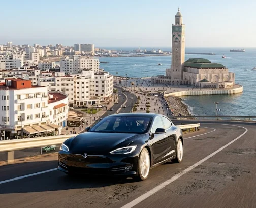 A black Tesla Model S drives on a Moroccan coastal road towards the famous Hassan II Mosque in Casablanca.