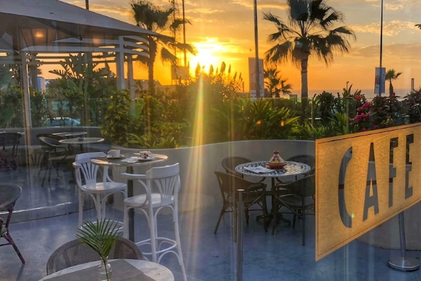 Vue depuis l'intérieur, à travers une vitre portant l'inscription "CAFÉ", d'une terrasse de café à Casablanca au coucher du soleil, avec tables, chaises blanches et sombres, palmiers et le ciel orange.
