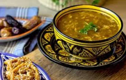 Un bol en céramique traditionnelle illustrant la gastronomie marocaine avec une soupe Harira fumante, accompagnée d'un gâteau Chebakia croustillant au sésame et d'une assiette de dattes.