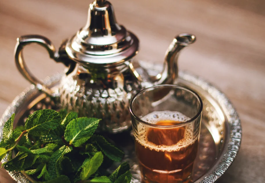 Un plateau en argent ciselé illustrant la gastronomie marocaine avec une théière en métal, un verre de thé à la menthe chaud bien moussé et un bouquet de feuilles de menthe fraîche.