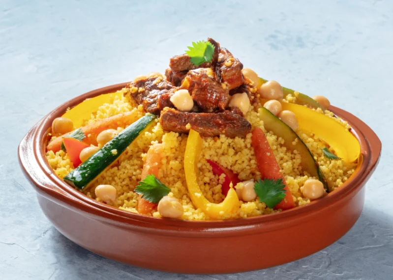 A vibrant ceramic bowl displaying Moroccan Gastronomy with a mountain of steamed couscous grains, topped with tender beef pieces, chickpeas, carrots, zucchini, and yellow bell peppers.