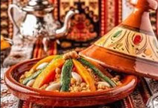 Couscous marocain traditionnel aux sept légumes présenté dans un plat en terre cuite avec une théière en arrière-plan.