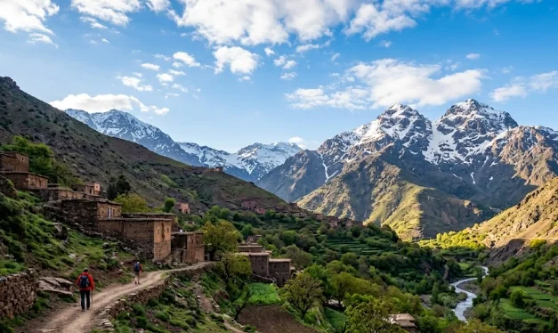 Deux randonneurs marchant sur un sentier dans les montagnes de l'Atlas, entourés de maisons traditionnelles berbères en argile, de vallées vertes en terrasses, d'une rivière et de sommets enneigés au Maroc.